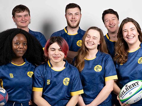 Seven students in ARU sports kit posing for a studio photo, two holding rugby balls