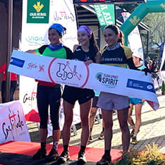 Three women in running clothes standing at a finish line with a banner