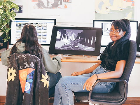 Two students, one facing the camera, working on digital art on computer screens