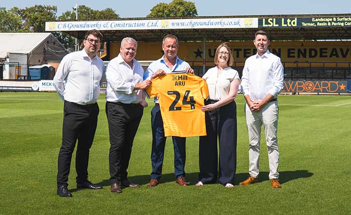 Representatives from ARU and Cambridge United standing on the pitch with an ARU-branded Cambridge United football shirt