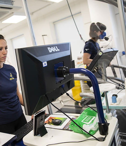 A student on a computer while a student behind on a treadmill wears an oxygen mask