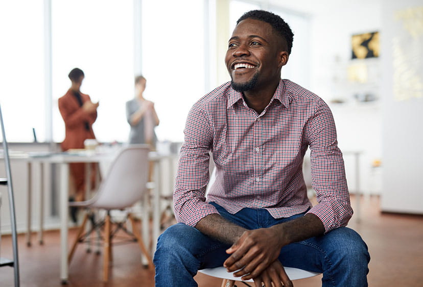 A smiling man sitting on a chair in a meeting room with two colleagues in the background