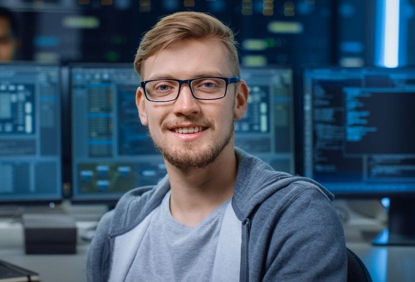 A man with glasses sits in front of a computer screen showing data and smiles at the camera