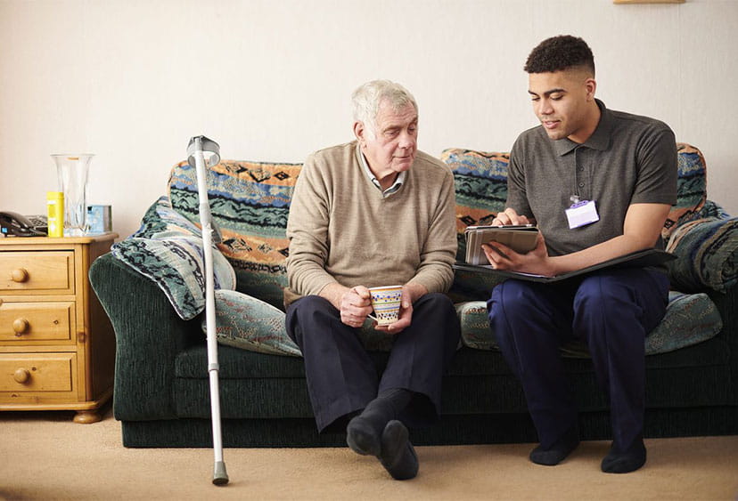 A carer sitting next to an elderly person on a sofa, showing them something on a tablet