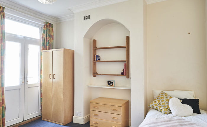 A view of a sparse bedroom showing bed, chest, wardrobe, shelf and windows.  