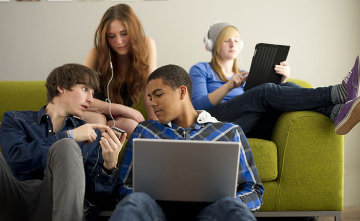 Four teenagers using tablets, phones and laptops, two sitting on a green sofa, and two sitting in front of it