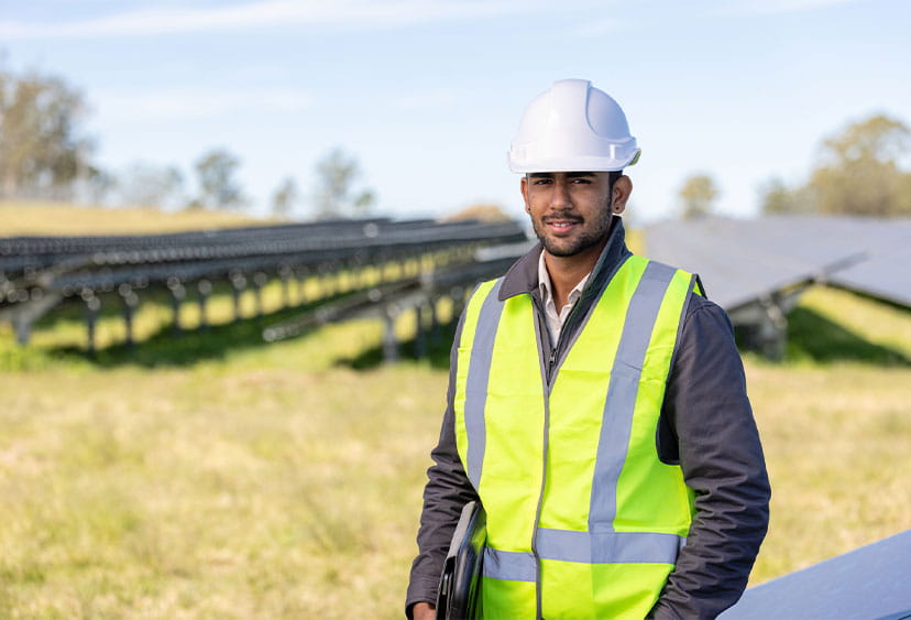 Male student in hi-vis jacket and hard hat stood next to solar panels