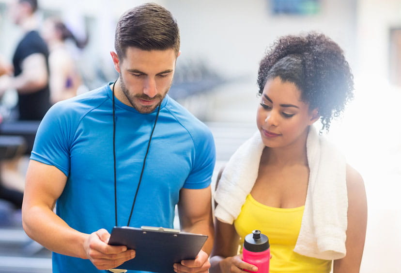 A woman dressed for a workout with a water bottle and towel, and a male fitness instructor looking at a clipboard together in a gym