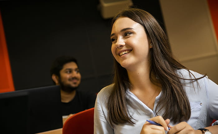 A smiling student holding a pen with another student using a computer in the background