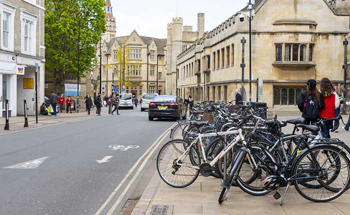 Cambridge street scene featuring a crowd of parked bicycles, a road with a cycle lane, pedestrians, and cars and historical buildings in the distance