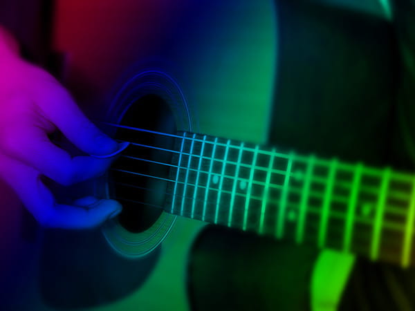 A hand plucking guitar strings with a plectrum, lit in shades of purple, blue and green