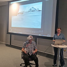 Neil Marsden using a VR headset, assisted by Lesley Johnston, in a lecture hall