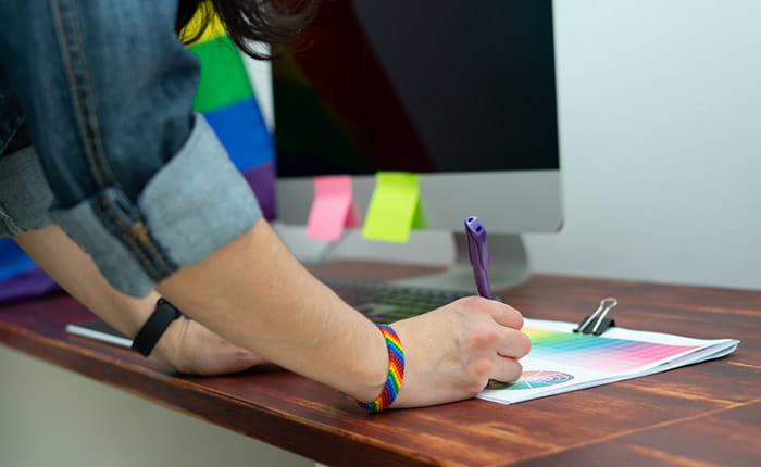 Close-up of an arm and hand writing on a document in an office setting. The person is wearing a rainbow wristband and there's a rainbow flag behind their computer monitor
