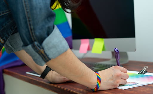 Close-up of an arm and hand writing on a document in an office setting. The person is wearing a rainbow wristband and there's a rainbow flag behind their computer monitor