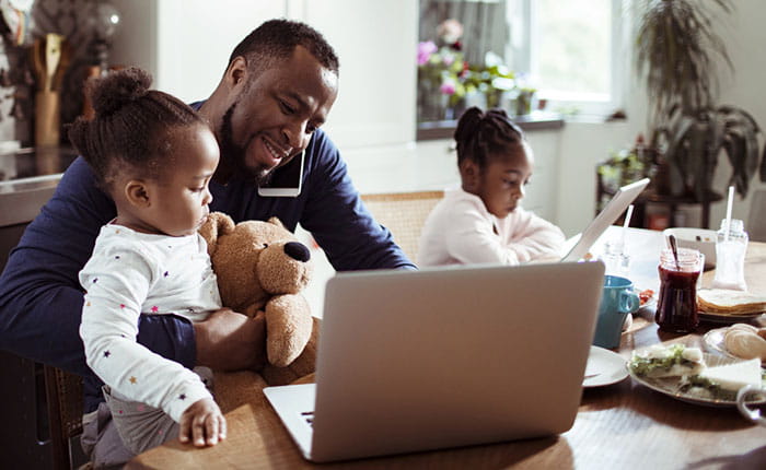 Someone working on a laptop at a kitchen table while on the phone, while cuddling a child, with another child next to them. 