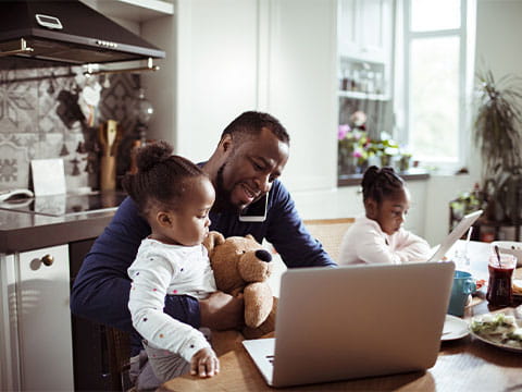 Someone working on a laptop at a kitchen table while on the phone, while cuddling a child, with another child next to them. 