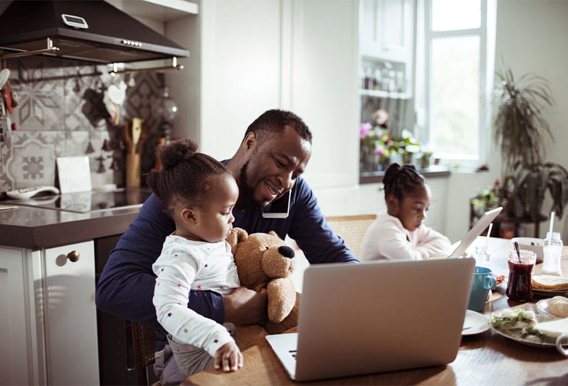 Someone working on a laptop at a kitchen table while on the phone, while cuddling a child, with another child next to them. 