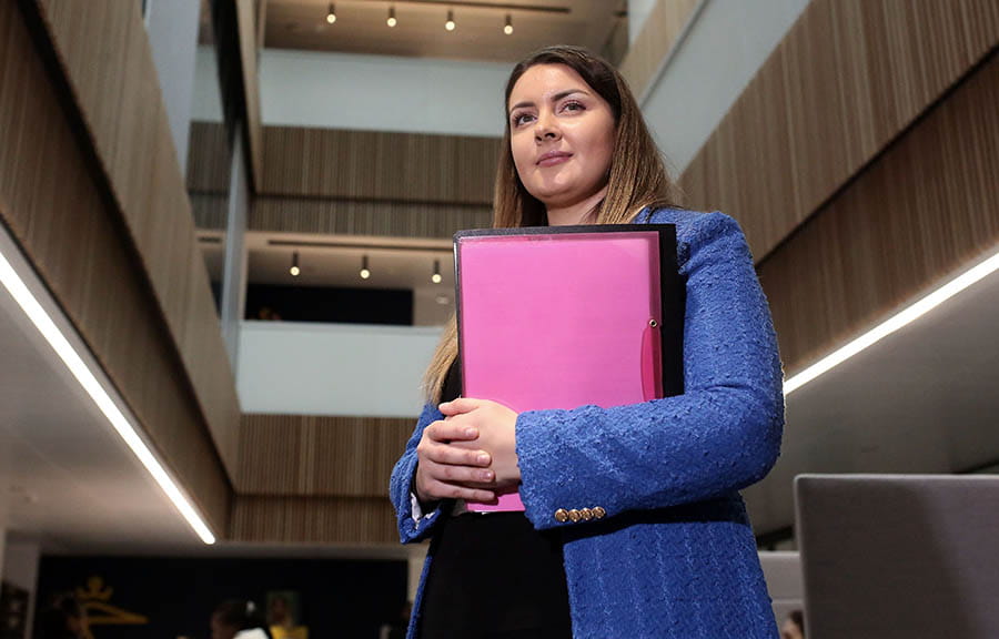 A student standing, holding a folder, in the main area of University House, ARU Peterborough