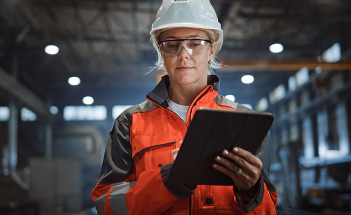A young woman in a white hard hat and orange hi viz jacket holding a tablet in a warehouse