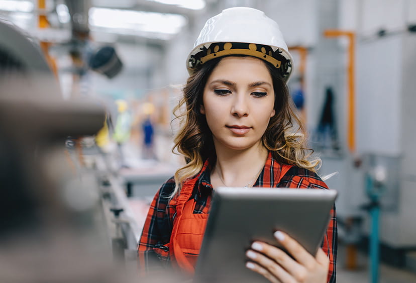 A young woman in a workshop, wearing a hard hat and using a tablet