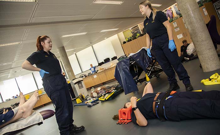 Two Paramedic Science students standing in the skills lab, with another lying on the floor in a head immobiliser