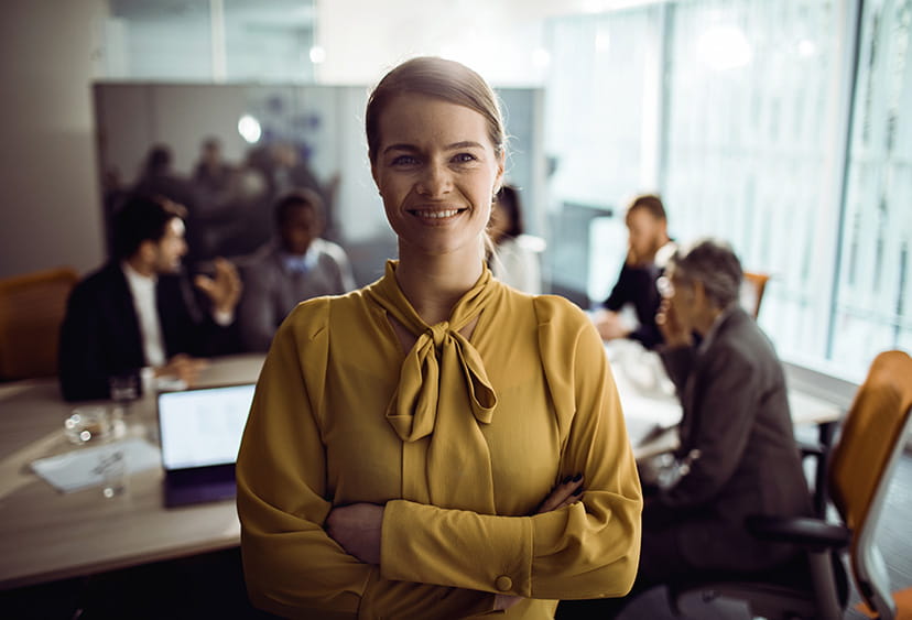 A smartly-dressed woman smiling at the camera, with colleagues meeting in the background