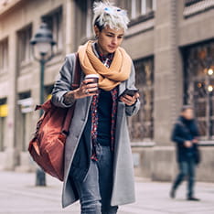 A young woman holding a takeaway cup of coffee and looking at her phone while walking down a city street