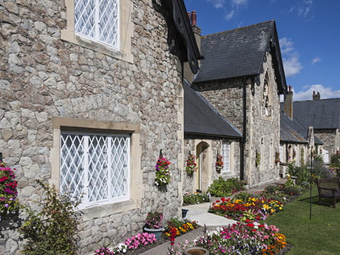 A row of stone almshouses fronted by flowerbeds, grass and a bench