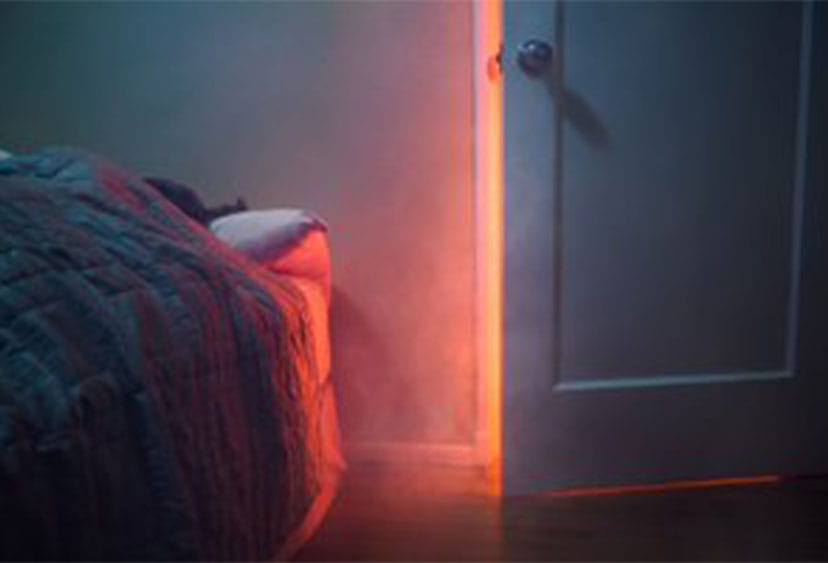Smoke coming into a bedroom though an ajar door with the glow of flames behind it