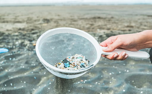 A hand holding a sieve containing microplastics above the sea shore