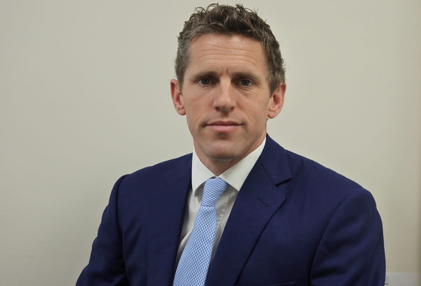 Headshot of Rupert Bourne. He has short, light brown hair and is wearing a dark blue suit jacket, white shirt, and light blue tie.