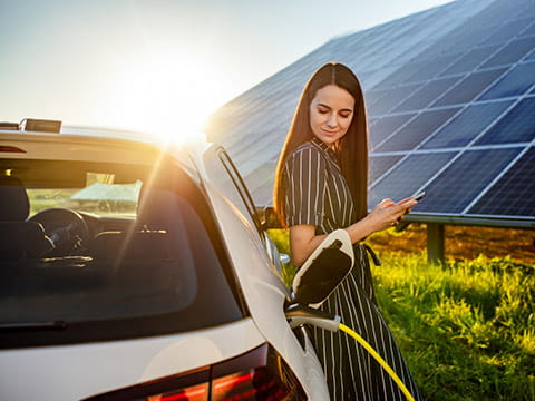 A person looking at their phone while charging an electric car near a large structure of solar panels