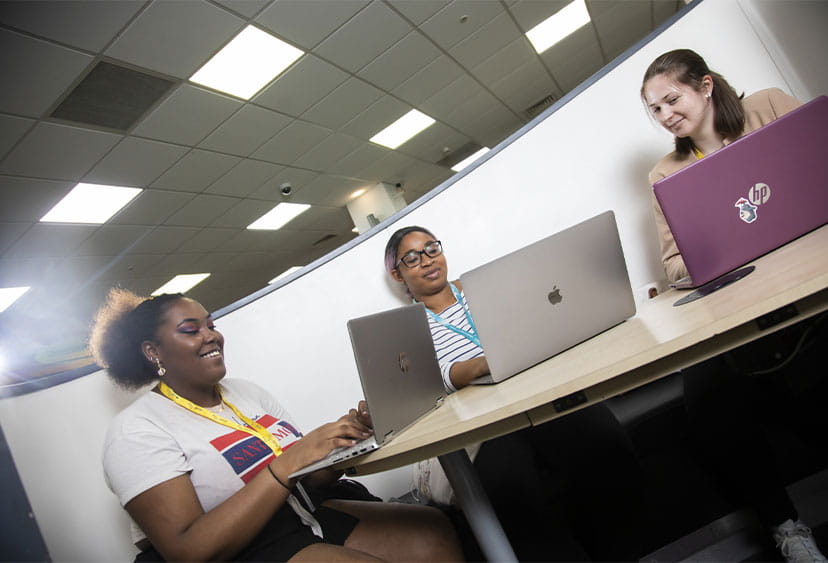 Three students at laptops in a cubicle, smiling together
