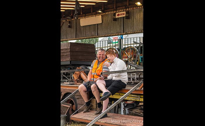 Two showmen laughing together on a trailer. One is older and wearing orange hi-viz over a grey t-shirt and shorts, the other is younger and wearing a white long-sleeved top and black shorts.