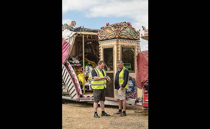 Two showmen in hi viz talking next to a packed-up carousel in a field