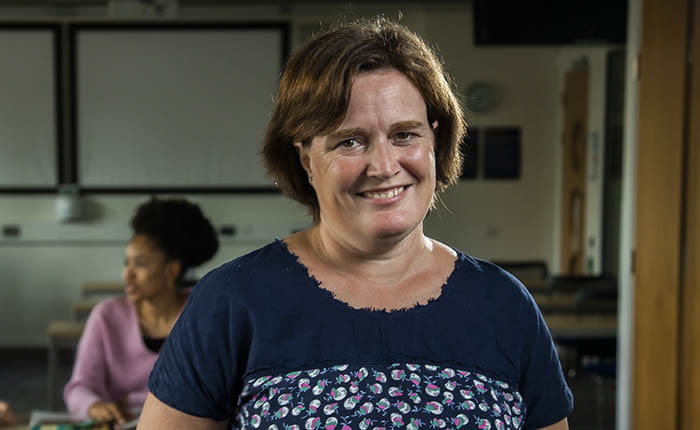 A Social Work student standing and smiling for the camera in a classroom, with a second student talking at a table in the background