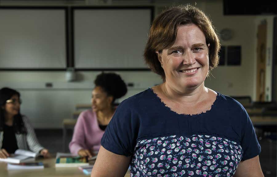 A Social Work student standing and smiling for the camera in a classroom, with two others talking at a table in the background