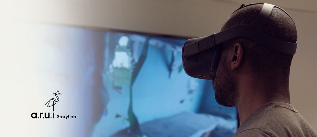 A man wearing a VR headset in front of a screen displaying blue graphics, with the ARU StoryLab logo superimposed