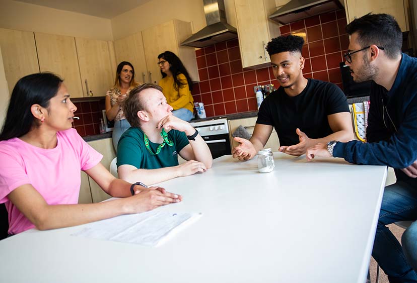 Six students, four sitting at a table and two standing, talking in a kitchen in university halls