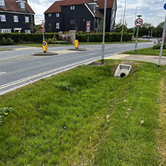 View of a main road with houses on the far side, and a grass swale with gabion wall on the near side