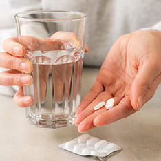 Close up of a pair of hands, one holding a glass of water, and the other holding two pills. The rest of the blister pack is on the surface in front of them