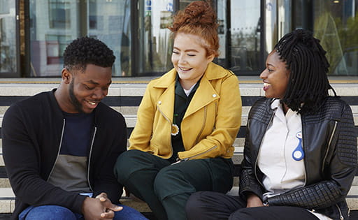Three students sitting on the steps in front of the revolving doors leading into ARU's School of Medicine on the Chelmsford campus