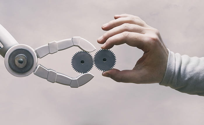 A robot claw and a human hand, both holding up cogs of the same size