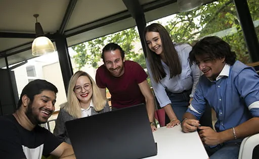 Five smiling students gathered around a laptop